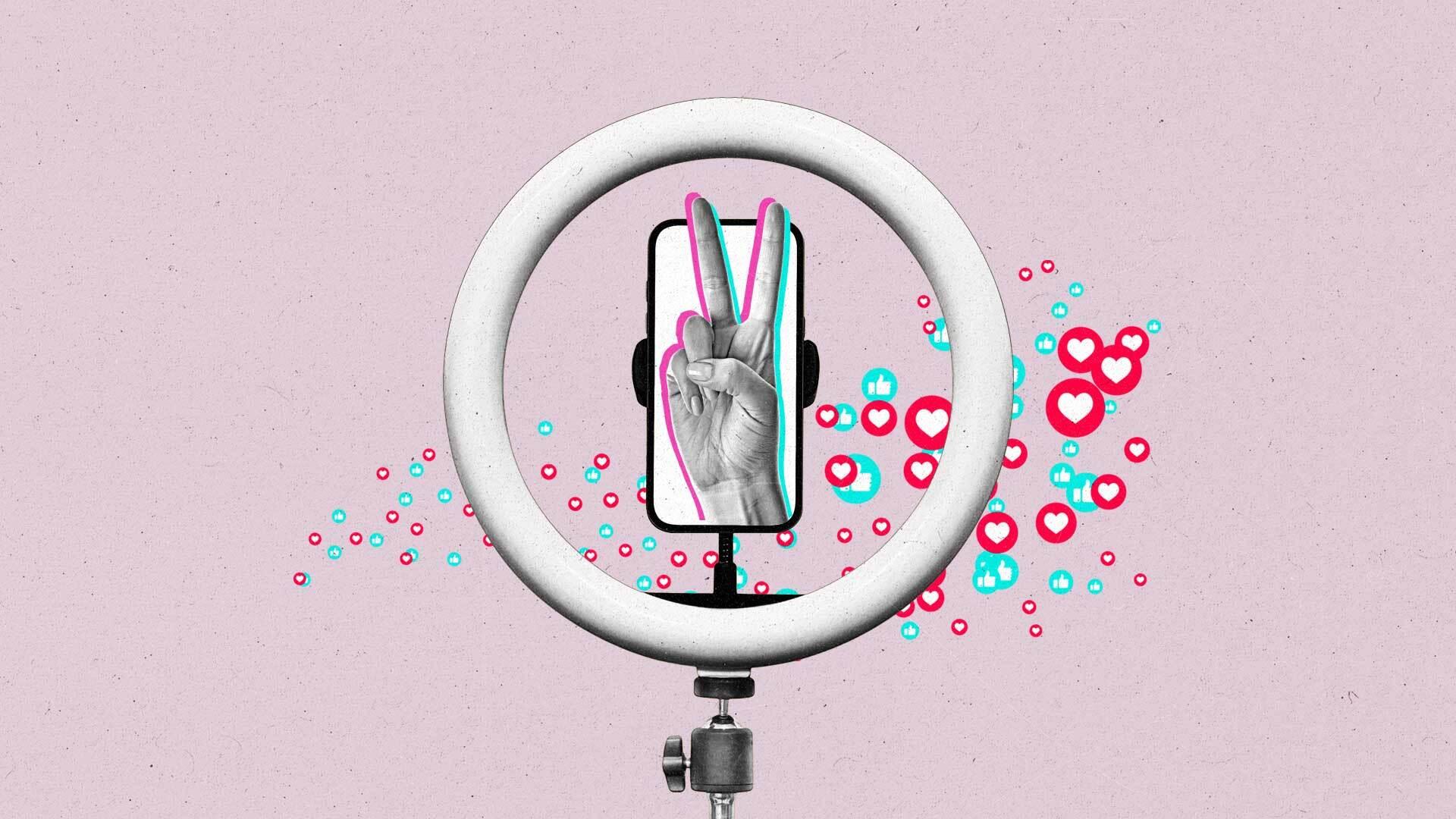 A hand making a peace sign sticks out of a smartphone within a ring light as like and love emoji float across it.